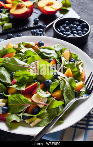 Nahaufnahme der Salat mit frischen Mangold Blätter, Pfirsich, Blaubeeren, Stücke von blauen Schimmel Käse serviert auf einem weißen Teller auf einen konkreten Tisch mit Zutaten Stockfoto