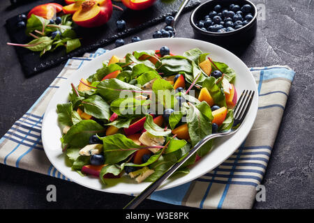 Nahaufnahme der Salat mit frischen Mangold Blätter, Pfirsich, Blaubeeren, Stücke von blauen Schimmel Käse serviert auf einem weißen Teller auf einen konkreten Tisch mit Zutaten Stockfoto