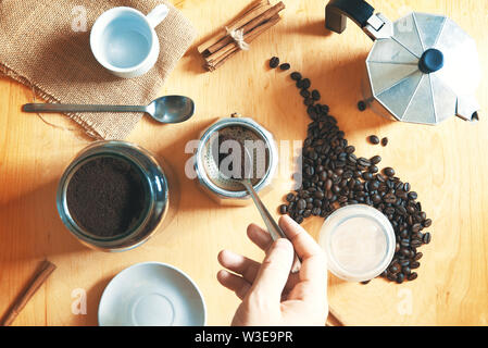 Hand mit einem Löffel Vorbereitung einige Expresso Café mit Kaffee Topf auf einem rustikalen Holztisch. Nächste, gemahlenen Kaffee und Bohnen mit Zimt. Stockfoto