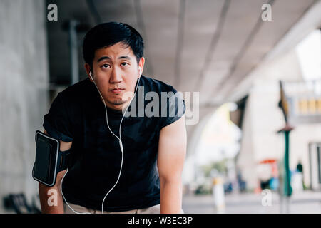 Erschöpft Asiatischen männlich Musik hören und lehnte sich auf Knien beim Stehen auf unscharfen Hintergrund der Stadt Straße in Outdoor Training Stockfoto