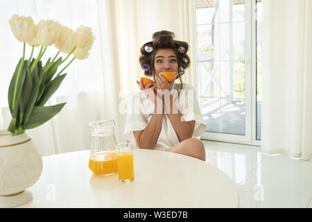 Schöne junge brünette Frau in Lockenstäbe beißen orange und orange in einem anderen Hand in helle Küche Stockfoto