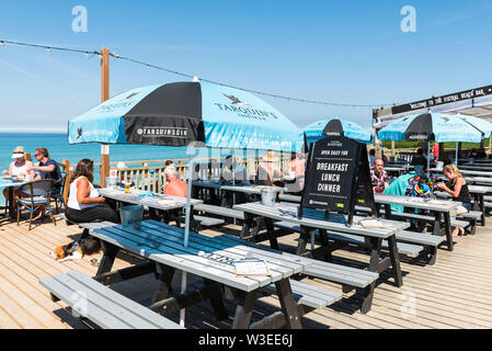 Urlauber entspannen und genießen Sie die Sonne auf dem Deck der Fistral Beach Bar in Newquay in Cornwall. Stockfoto