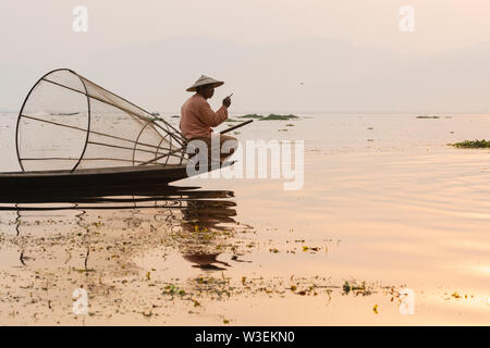 Inle, Myanmar - März 2019: Traditionelle Burmesische Bein rudernden Fischer am Inle See bei Sonnenaufgang. Das Sitzen auf dem Boot und Rauchen. Stockfoto