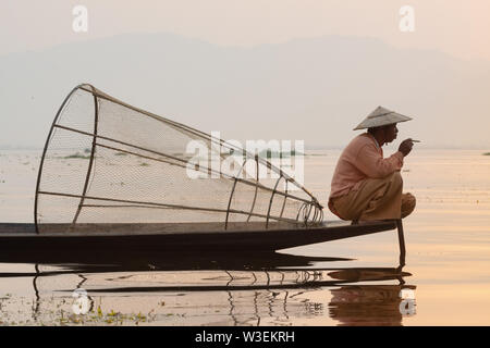 Inle, Myanmar - März 2019: Traditionelle Burmesische Bein rudernden Fischer am Inle See bei Sonnenaufgang. Das Sitzen auf dem Boot und Rauchen. Stockfoto