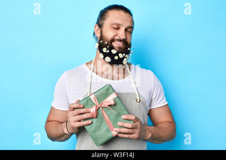 Happy positive stattlicher Mann mit Blumen in seinem Bart holding Geschenk Box in die Hände und schauen in die Kamera. Isolierte blauen Hintergrund. Lieferung club, Ma Stockfoto