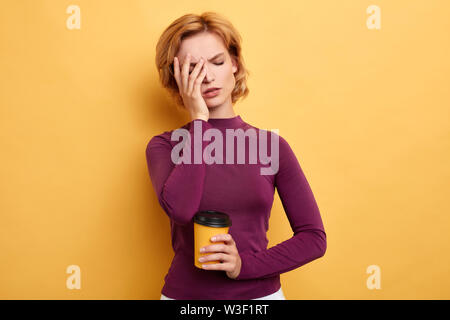 Müde, schläfrig, schöne Frau deckt die Hälfte der Fläche mit Palm, hat traurige Ausdruck, schließt die Augen, halten Einweg Becher mit Tee am Morgen, isoliert Stockfoto