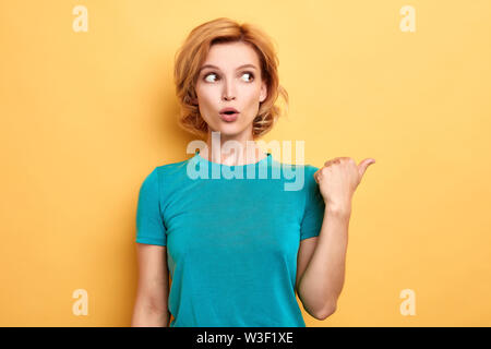 Erstaunt emotionale blonde Frau hat verwundert und erschrockenen Ausdruck, sieht überraschend beiseite, zeigt mit Daumen. Bis portrait schließen, isoliert Stockfoto