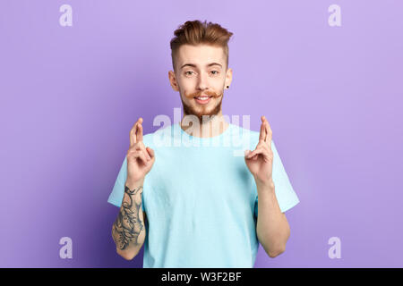 Hübscher junger Mann mit lustigen Schnurrbart und Tattoo steht vor blauem Hintergrund mit gekreuzten Fingern, und wenn man die Kamera bitten um gute Luc Stockfoto