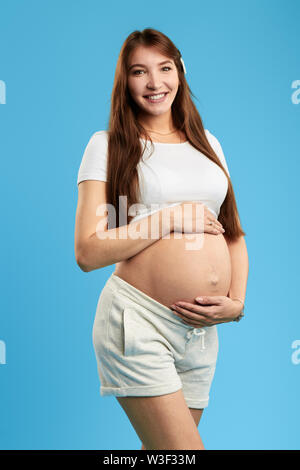Positive schöne junge Frau mit großen Bauch in die Kamera schaut. Glück, neues Leben, isoliert blauen Hintergrund. studio Shot, erben und Nachfolger Stockfoto