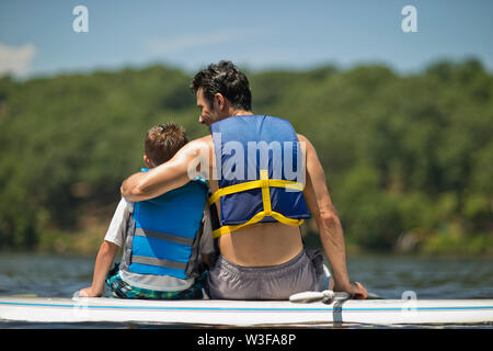 Vater mit seinen Arm um seinen Sohn, da sie auf einem paddleboard sitzen. Stockfoto