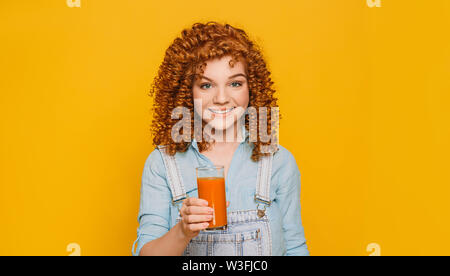 Curly rothaarige Frau mit einem Glas frischen Karottensaft auf gelbem Hintergrund. Versuchen Sie, eine gesunde Vitamin Saft Stockfoto