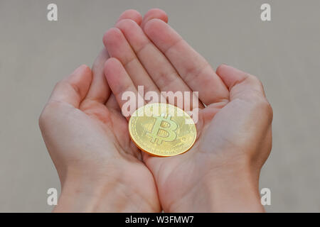 Eine goldene cryptocurrency bitcoin Münze auf die längliche Frauen Hände auf einem grauen Hintergrund. Eine Frauen Hände halten einen Großen goldenen Medaille der bitcoin. Stockfoto