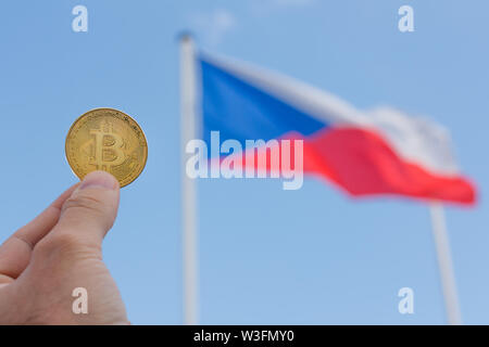 Ein Mann die Finger halten einen Großen goldenen Medaille der bitcoin vor der Tschechischen Republik Flagge und blauer Himmel. Es gibt einen großen goldenen bitcoin Münze auf den Tschechischen Stockfoto