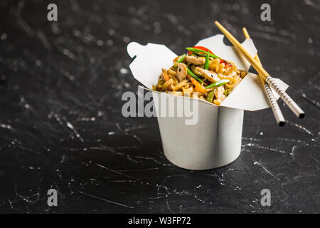 Chinesische Nudeln in einer Box mit Hähnchen und Gemüse mit Stöcken. Wok Verpflegung aus Restaurant auf dunklem Hintergrund aus der Seite. Stockfoto