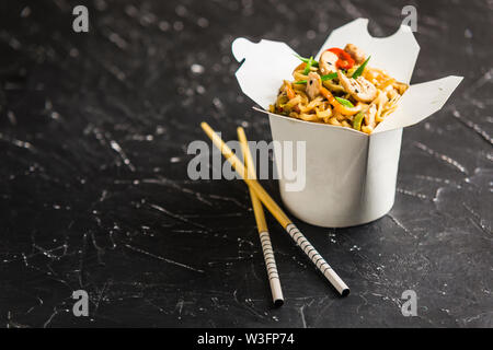 Chinesische Nudeln in einer Box mit Hähnchen und Gemüse mit Stöcken. Wok Verpflegung aus Restaurant auf dunklem Hintergrund aus der Seite. Stockfoto