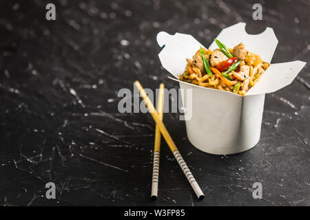 Chinesische Nudeln in einer Box mit Hähnchen und Gemüse mit Stöcken. Wok Verpflegung aus Restaurant auf dunklem Hintergrund aus der Seite. Stockfoto