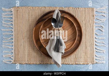 Ort, Einstellung auf Glasgewebe Tischset auf blauem Hintergrund mit zwei gestapelten Holzplatten und Bettwäsche Serviette mit Black Metal Bestecke und Korbwaren Serviette r Stockfoto