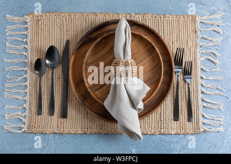 Ort, Einstellung auf Glasgewebe Tischset auf blauem Hintergrund mit zwei gestapelten Holzplatten und Bettwäsche Serviette mit Black Metal Bestecke und Korbwaren Serviette r Stockfoto