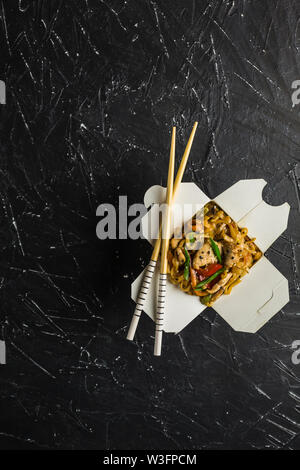 Chinesische Nudeln in einer Box mit Hähnchen und Gemüse mit Stöcken. Wok Verpflegung aus Restaurant auf dunklem Hintergrund von oben ansehen. Stockfoto