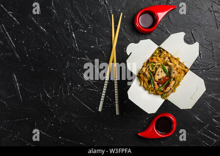 Chinesische Nudeln in einer Box mit Hähnchen und Gemüse mit Stöcken. Wok Verpflegung aus Restaurant auf dunklem Hintergrund von oben ansehen. Stockfoto