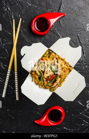 Chinesische Nudeln in einer Box mit Hähnchen und Gemüse mit Stöcken. Wok Verpflegung aus Restaurant auf dunklem Hintergrund von oben ansehen. Stockfoto