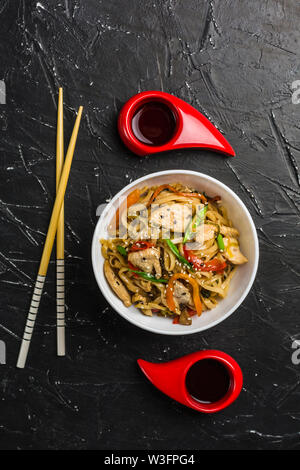 Chinesische Nudeln in eine Schale mit Hähnchen und Gemüse mit Stöcken. Wok Essen aus dem Restaurant auf einem dunklen Hintergrund von oben gesehen. Stockfoto