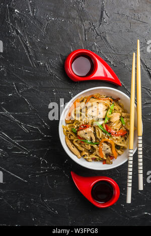 Chinesische Nudeln in eine Schale mit Hähnchen und Gemüse mit Stöcken. Wok Essen aus dem Restaurant auf einem dunklen Hintergrund von oben gesehen. Stockfoto