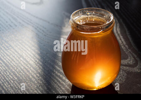 Delicious Organic Natural Bio Honig in einem offenen Glas Glas auf Holztisch mit Reflexion der Sonne, schwarzer Hintergrund, Nahaufnahme Stockfoto