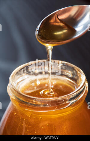 Delicious Organic Natural Bio Honig gießt der Löffel in ein Glas Glas auf Holztisch mit Reflexion der Sonne, schwarzer Hintergrund, vertikal Stockfoto