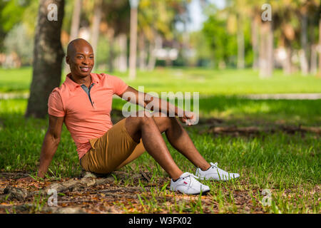 Foto eines jungen schwarzen Mann im Park sitzen. Person legere pink Polo Shirt und Shorts Stockfoto