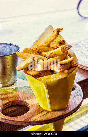 Pommes Frites Kartoffeln geschnitten in einem gelben Papiertüte, bereit zu essen, geschnitten in einer Holz- Halter in einer niederländischen Cafe geputtet. Holland Stockfoto