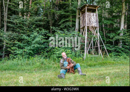 Old Hunter sitzt im Gras vor der seine Jagd Kanzel und liebevoll hält seine Irischen Setter Welpe in seinen Händen. Stockfoto