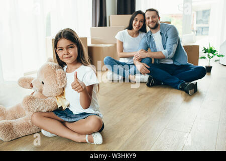 Kleines Mädchen Daumen oben, sitzen am Boden. Tochter Umzug in neues Haus mit Ihrer Familie Stockfoto