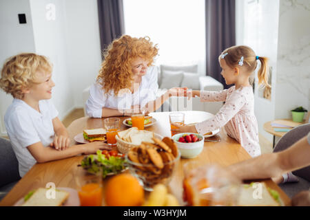 Glückliche Mutterschaft. Positive rothaarige Frau, die ihre Töchter Hand, während am Esstisch sitzen Stockfoto