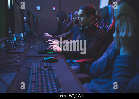 Seitenansicht von Konzentriertem süchtig multi-ethnischen Spieler sitzen in dunklen Raum der Computer Club und mit leistungsfähigen Computern für Online-Spiel Stockfoto