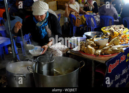 Die Gäste sitzen auf Kunststoff Tische und Stühle, Huhn porridge im Freien restaurant Night Food Market Essen, Kessel von Lebensmitteln auf Kohleofen in kaltem Wetter Stockfoto