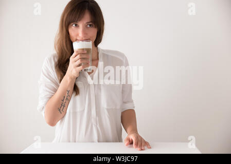 Junge brunette kaukasische Frau gegen weißen Hintergrund trinken Latte macchiato Kaffee und lächelnd Stockfoto