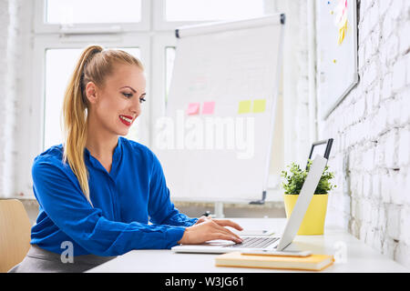 Wunderschöne lächelnden jungen Frau, die auf dem Laptop während in stilvollen, modernen Büro zu sitzen Stockfoto