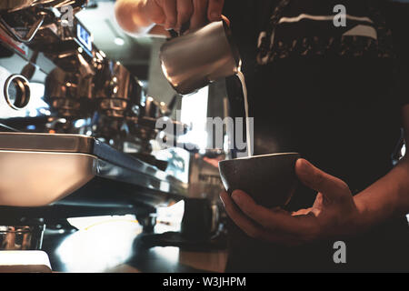 Barista Mann gießen Schlagsahne Milch aufschäumen Krug in der Tasse mit Kaffee stand vor der professionelle Kaffeemaschine im Cafe Stockfoto