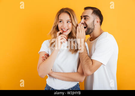 Bild der glücklichen Paare der Mann und die Frau in der Basic t-shirts whispering Geheimnisse oder Verleumder miteinander über Gelb Hintergrund isoliert Stockfoto