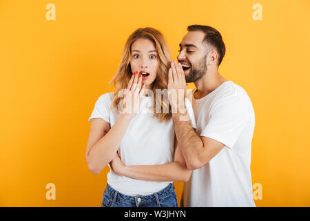 Bild von überrascht Paar der Mann und die Frau in der Basic t-shirts whispering Geheimnisse oder Verleumder miteinander über Gelb Hintergrund isoliert Stockfoto