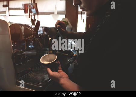 Barista Mann gießen Schlagsahne Milch aufschäumen Krug in der Tasse mit Kaffee trinken in der Nähe von professionelle Kaffeemaschine im Cafe Stockfoto