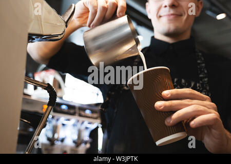 Lächelnd barista Mann gießen Schlagsahne Milch aufschäumen Krug in Pappbecher mit Kaffee stand vor der professionelle Kaffeemaschine im Cafe Stockfoto