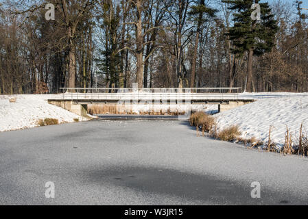 Gefrorener Mlynka Bach mit Brücke oben und Bäumen im Hintergrund auf Park Bozeny Nemcove öffentlichen Park in Karvina Stadt in der Tschechischen republik Stockfoto