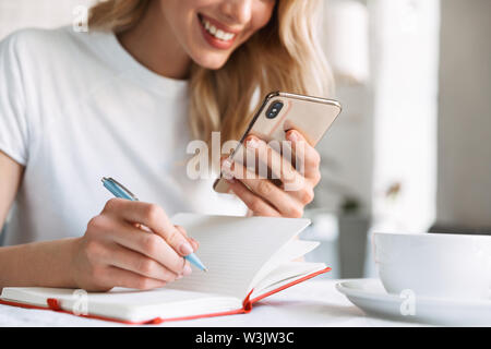 Zugeschnittenes Bild eines schönen jungen blonden Frau im Cafe in Innenräumen, über Handy, während sich Notizen Stockfoto