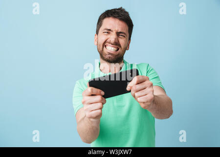 Bild einer aufgeregt emotionale Jungen gut aussehenden bärtigen Mann über blaue Wand Hintergrund spielen per Handy isoliert posieren. Stockfoto