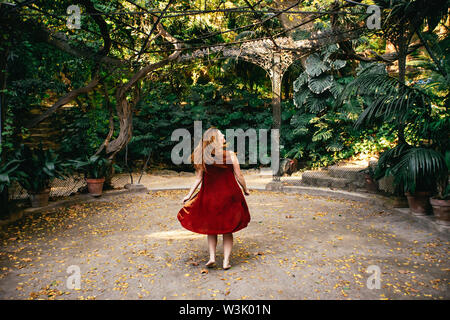 Glückliche Frau twirling Um in einem roten Kleid mit langen Haare hinter ihr fließt auf einen Fußweg im La Concepcion Historical-Botanical Garten, Malag Stockfoto