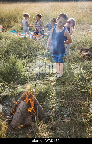 Little Boy rösten Marshmallow über einem Lagerfeuer. Familie Ausgabe Zeit zusammen auf der Wiese, in der Nähe der Natur. Eltern und Kinder sitzen auf einer Decke Stockfoto