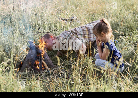Man beginnt ein Lagerfeuer, das Blasen auf ein Feuer. Kleines Mädchen sitzen im Gras neben einem Lagerfeuer. Ehrliche Menschen, echte Momente, in authentischen Situationen Stockfoto