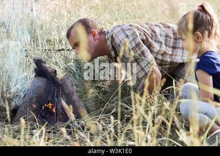 Man beginnt ein Lagerfeuer, das Blasen auf ein Feuer. Kleines Mädchen sitzen im Gras neben einem Lagerfeuer. Ehrliche Menschen, echte Momente, in authentischen Situationen Stockfoto